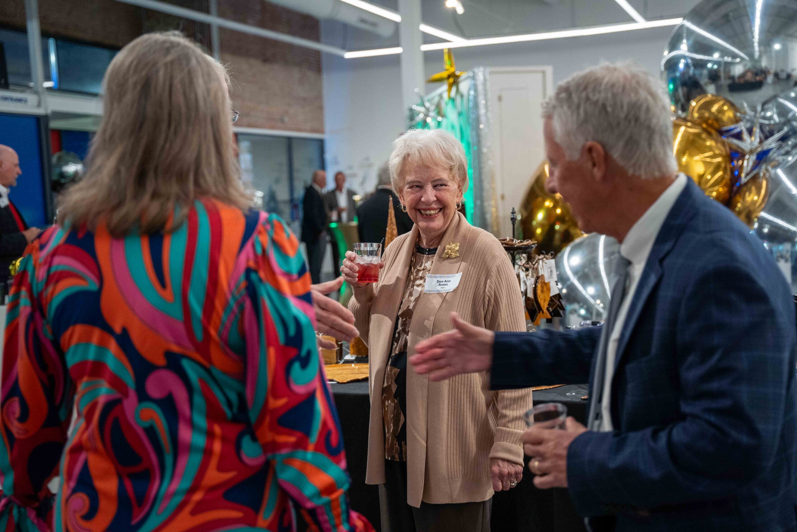 HOLIDAY INVITATIONAL 2025 an older woman stands holding a red mocktail smiling and talking to a gentleman in a suit holding out his hand